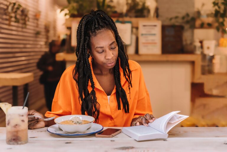an African-American woman reading a book while eating a dinner.